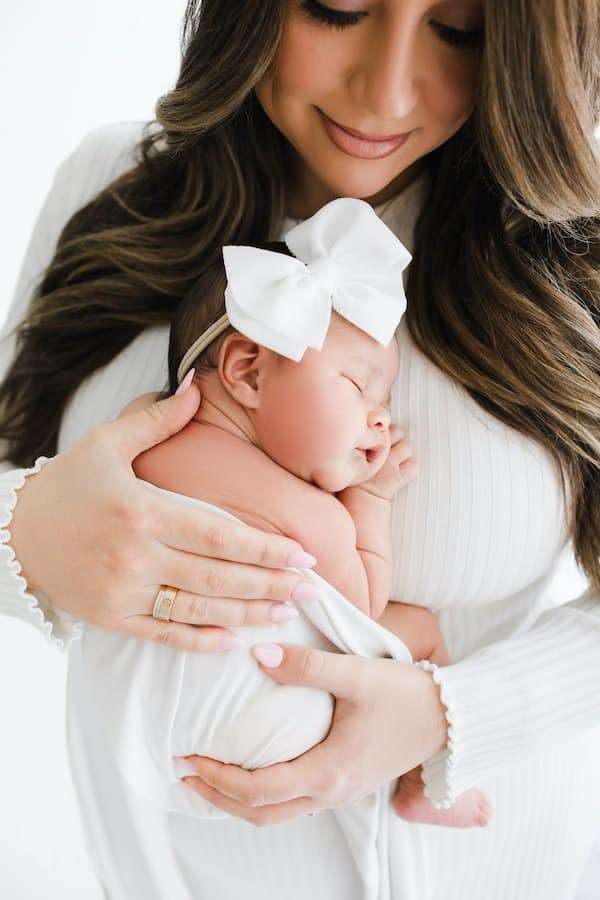 Woman in a white top holds a sleeping newborn wrapped in a white blanket, wearing a large white bow headband.