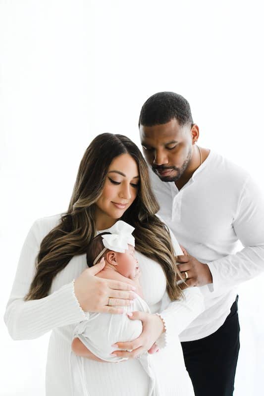 A woman holds a sleeping baby with a white bow while a man stands close beside them, all dressed in white against a bright background.