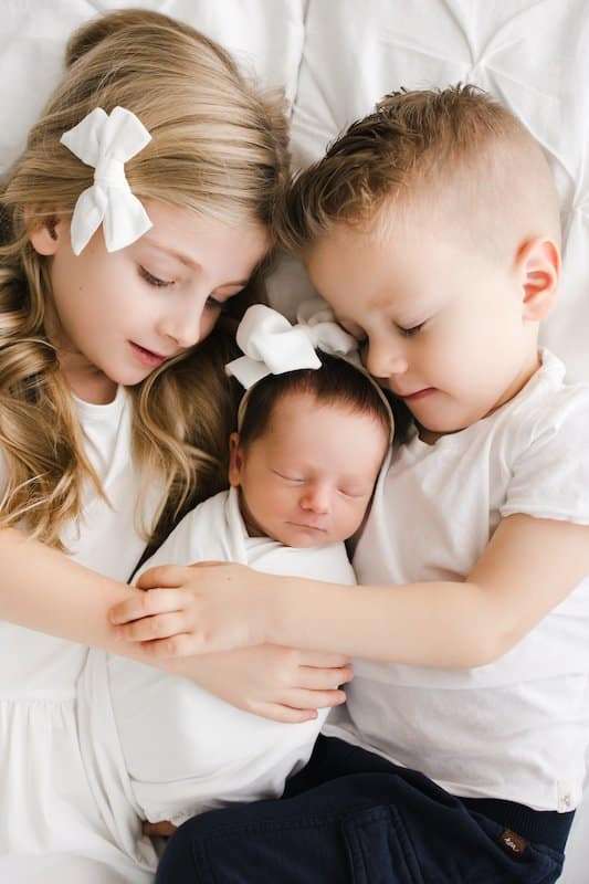A young girl and boy, both in white tops, gently cuddle a swaddled newborn baby lying between them on a white bed.