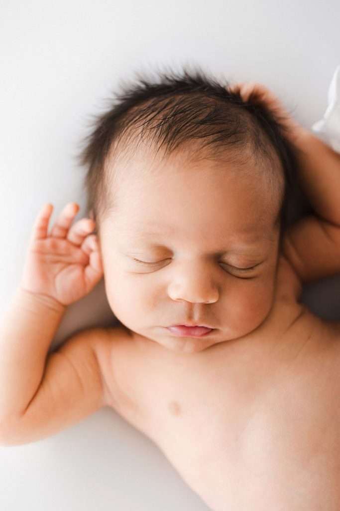 Newborn baby with closed eyes lying on a white surface, one arm raised above the head, and wearing no clothes.