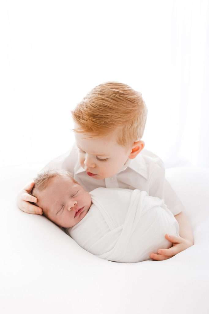 Young boy gently holds and looks down at a newborn baby wrapped in a white blanket, both dressed in white, against a white background.