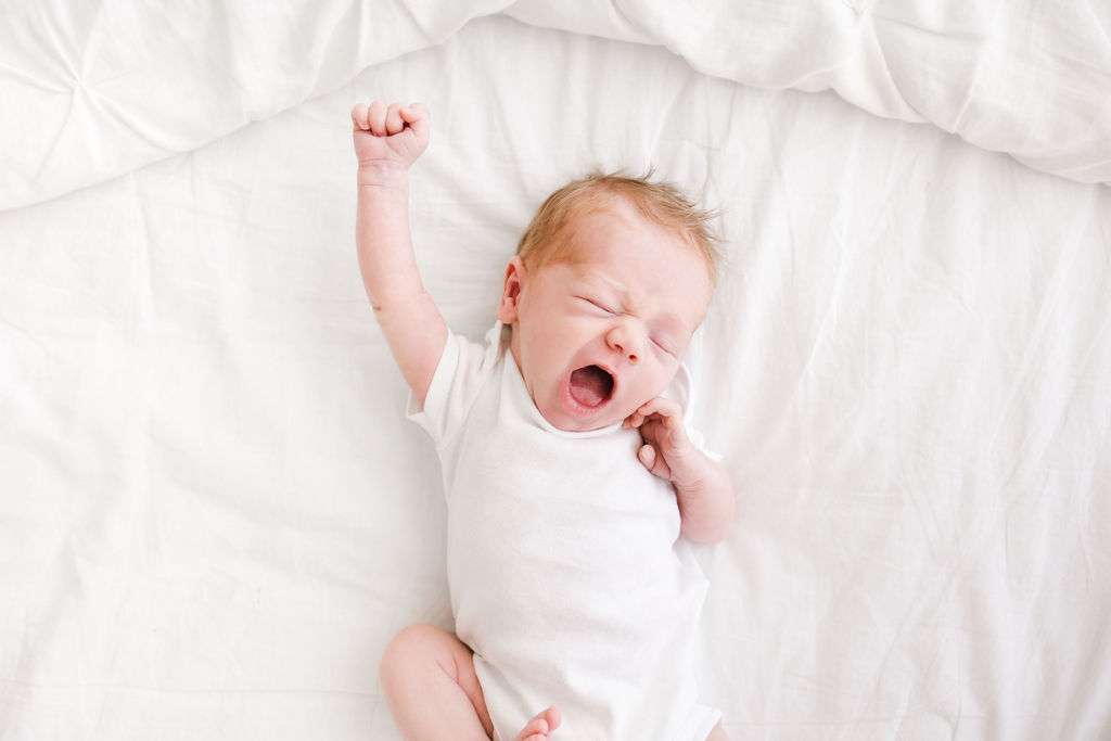 Newborn baby lying on a white bed, wearing a white onesie, yawning with one arm raised.