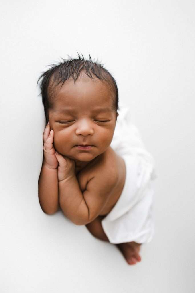 Newborn baby with closed eyes lies on a white surface, resting one cheek on their hand and wrapped in a white blanket.