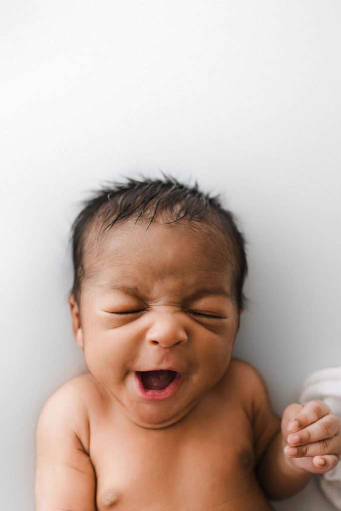 A newborn baby with closed eyes and mouth open, possibly yawning, against a plain white background.