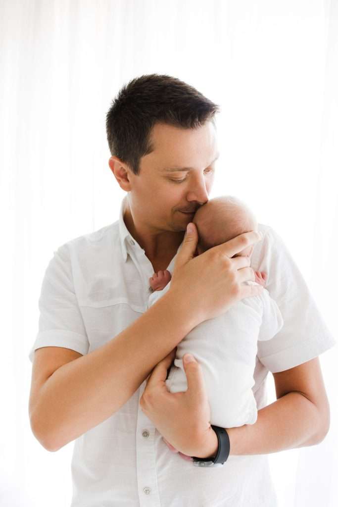 A man in a white shirt holds and gently kisses a newborn baby, both dressed in white, standing in front of a bright, softly lit background.