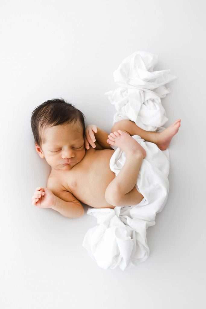 Newborn baby with dark hair lying on a white blanket, sleeping with arms and legs relaxed, photographed from above against a plain white background.