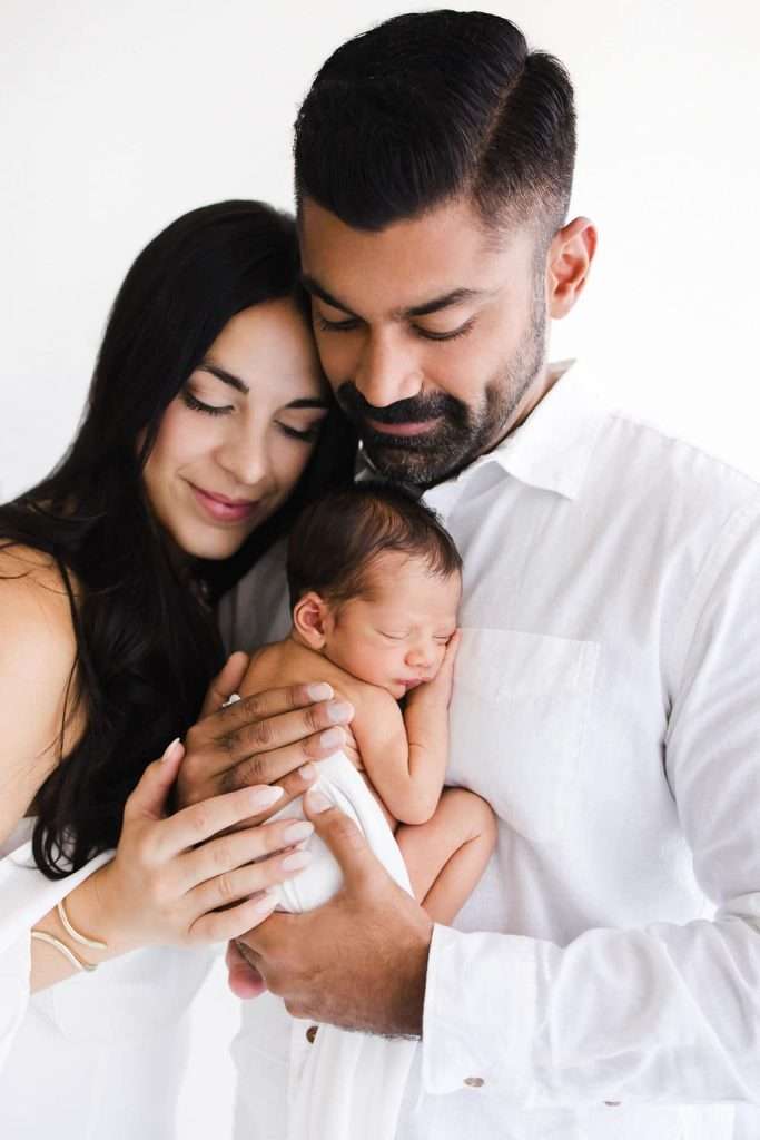 A couple dressed in white gently embraces a sleeping newborn baby, all three close together against a plain white background.