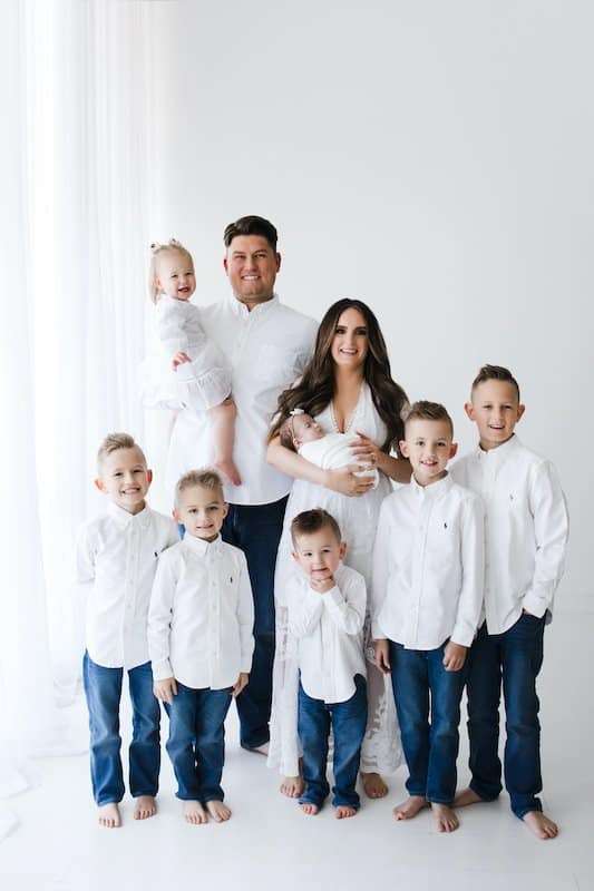 A family of eight poses together in a white studio. The parents stand at the back, holding two small children, with four boys standing in front. All wear white tops and blue jeans.
