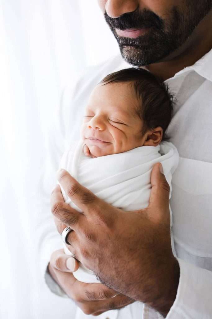 A man in a white shirt gently holds a swaddled newborn baby who is smiling with eyes closed.