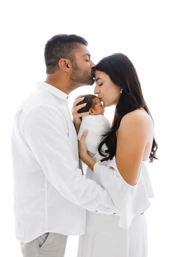 A man kisses a woman's forehead as she holds a newborn baby, all dressed in white, against a white background.