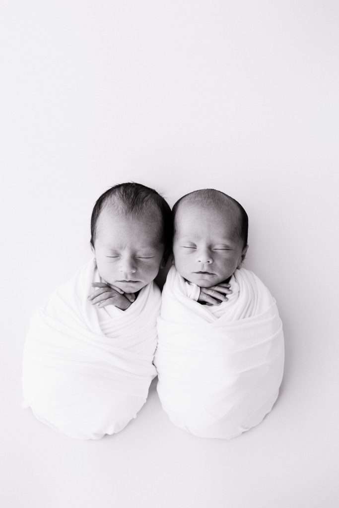 Two newborn babies are swaddled in white blankets and lying side by side on a white surface, both with their eyes closed.
