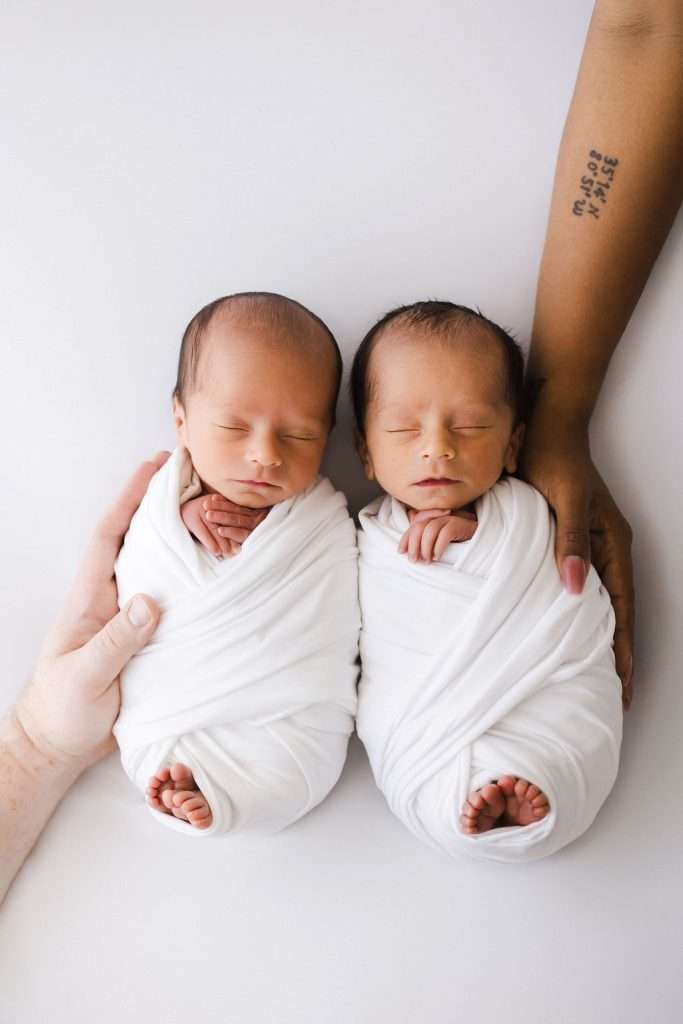 Two newborn babies swaddled in white blankets lie side by side, each gently held by an adult hand, one light-skinned and one dark-skinned, against a white background.