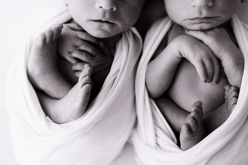 Black and white photo of two swaddled newborn babies lying side by side, with their feet and hands visible, wrapped in soft white blankets.