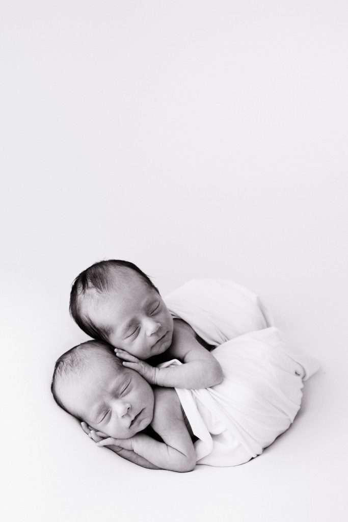 Two newborn babies wrapped in white cloth lie peacefully, one resting its head on the other’s back, against a plain background.