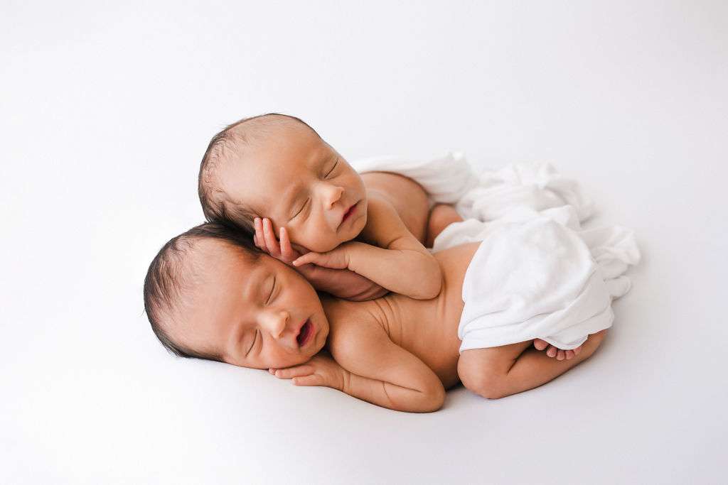 Two newborn babies sleep closely together on a white blanket, with one lying on top of the other. Both are unclothed except for white wraps covering their lower bodies.