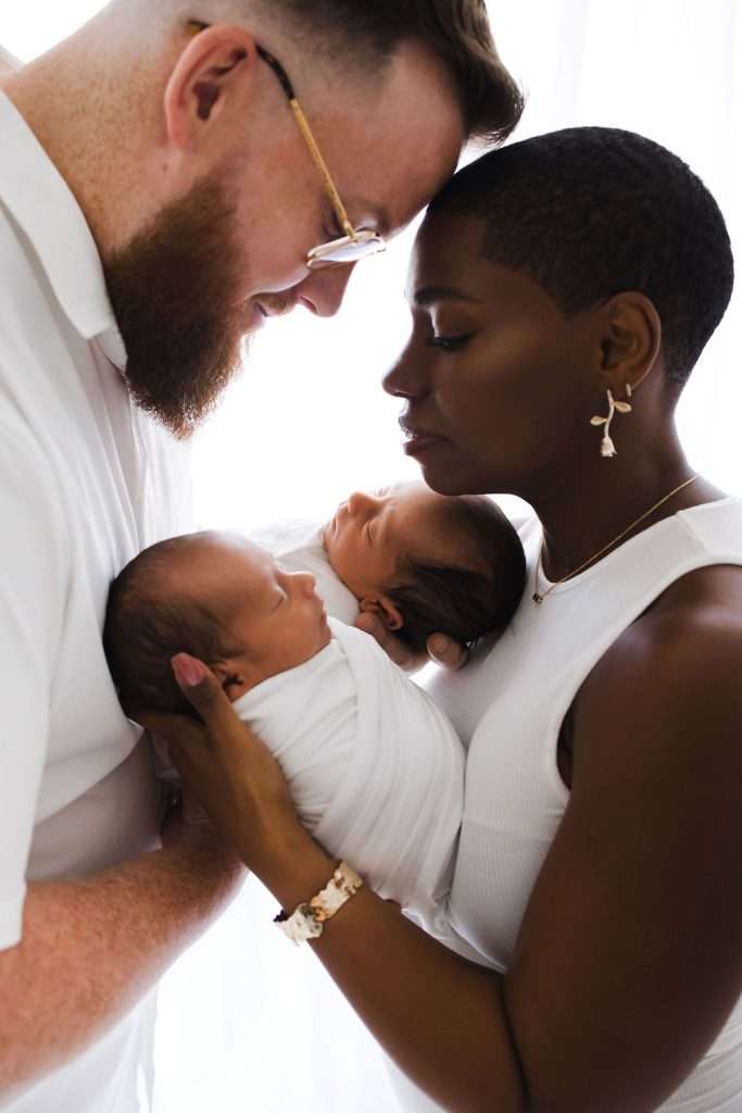 A couple stands close together, foreheads touching, each holding a swaddled newborn baby. They all wear white clothing against a bright, soft background.