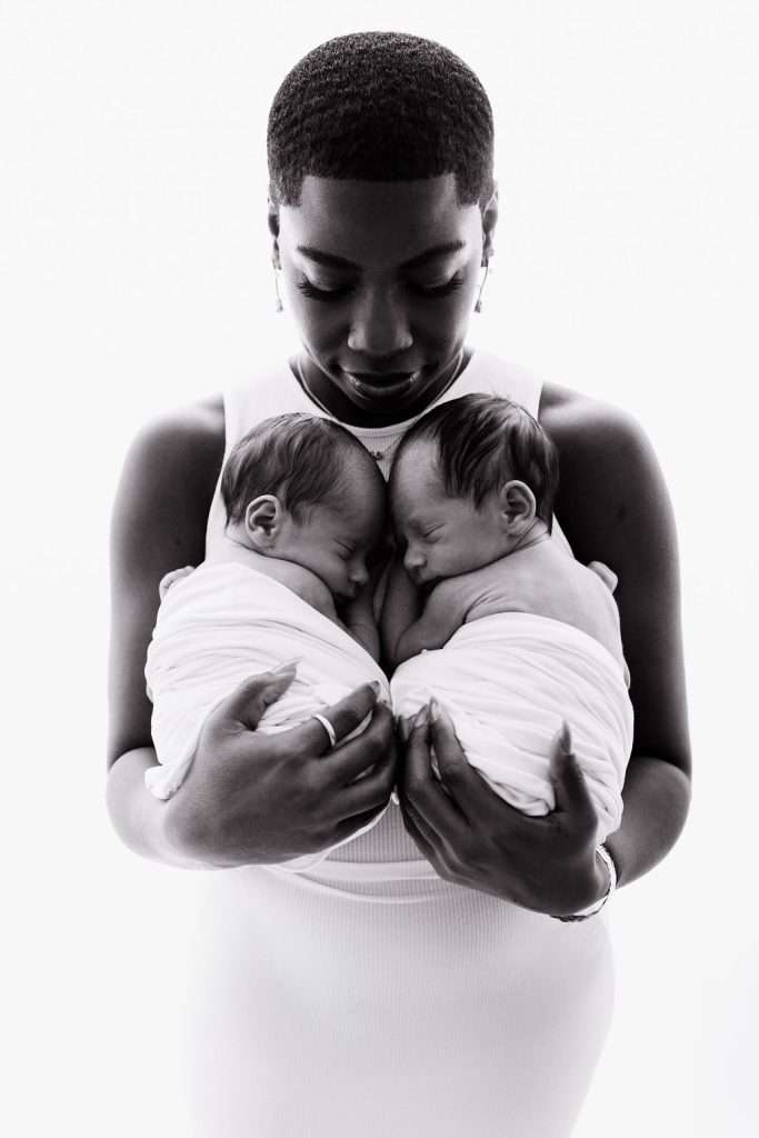 A woman in a white dress holds two swaddled newborns close to her chest, looking down at them affectionately against a white background.