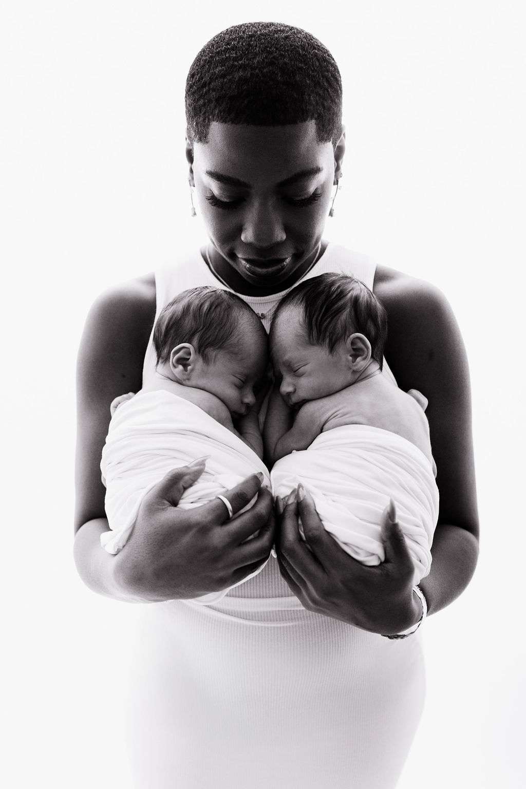 A woman in a white dress holds two swaddled newborns close to her chest, looking down at them affectionately against a white background.