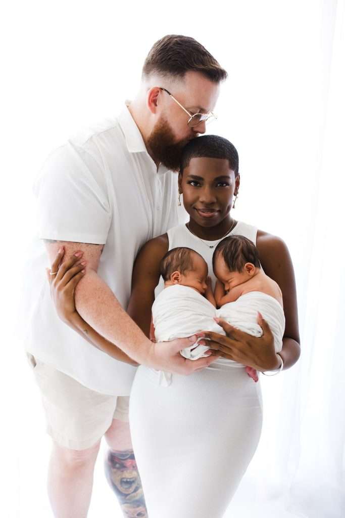 A couple stands together, the woman holding two swaddled newborns in her arms. All four wear white, posing against a bright, white background.