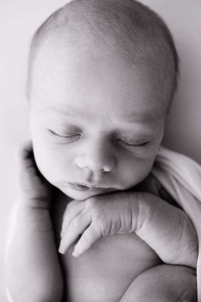 Black and white close-up photo of a sleeping newborn baby with eyes closed, resting their hand near their chin, wrapped in a light blanket.