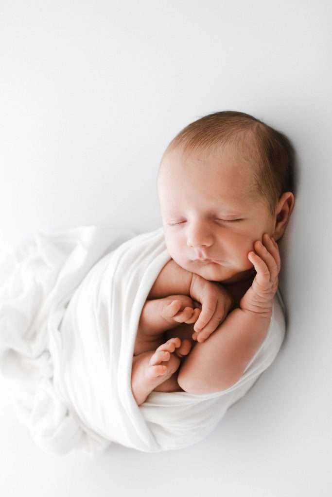 Newborn baby wrapped in a white blanket, sleeping on a white surface with hands and feet tucked close to the body.