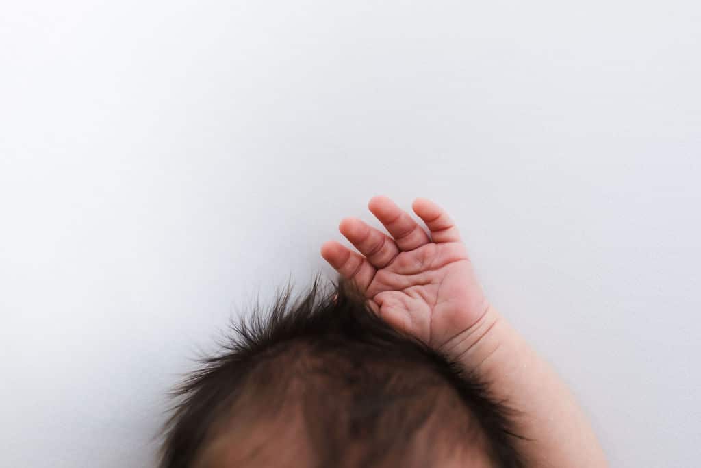 A close-up of a baby's head with dark hair and one hand raised against a plain white background.