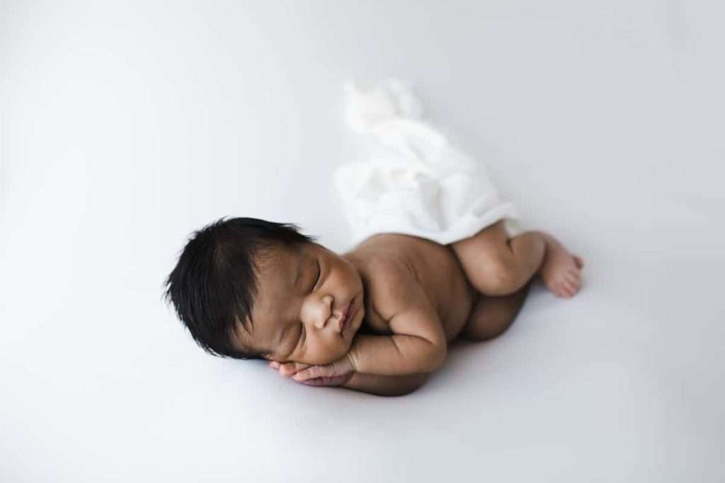 Newborn baby sleeping on a white surface with a white cloth partially covering their lower body, hands tucked under cheek.