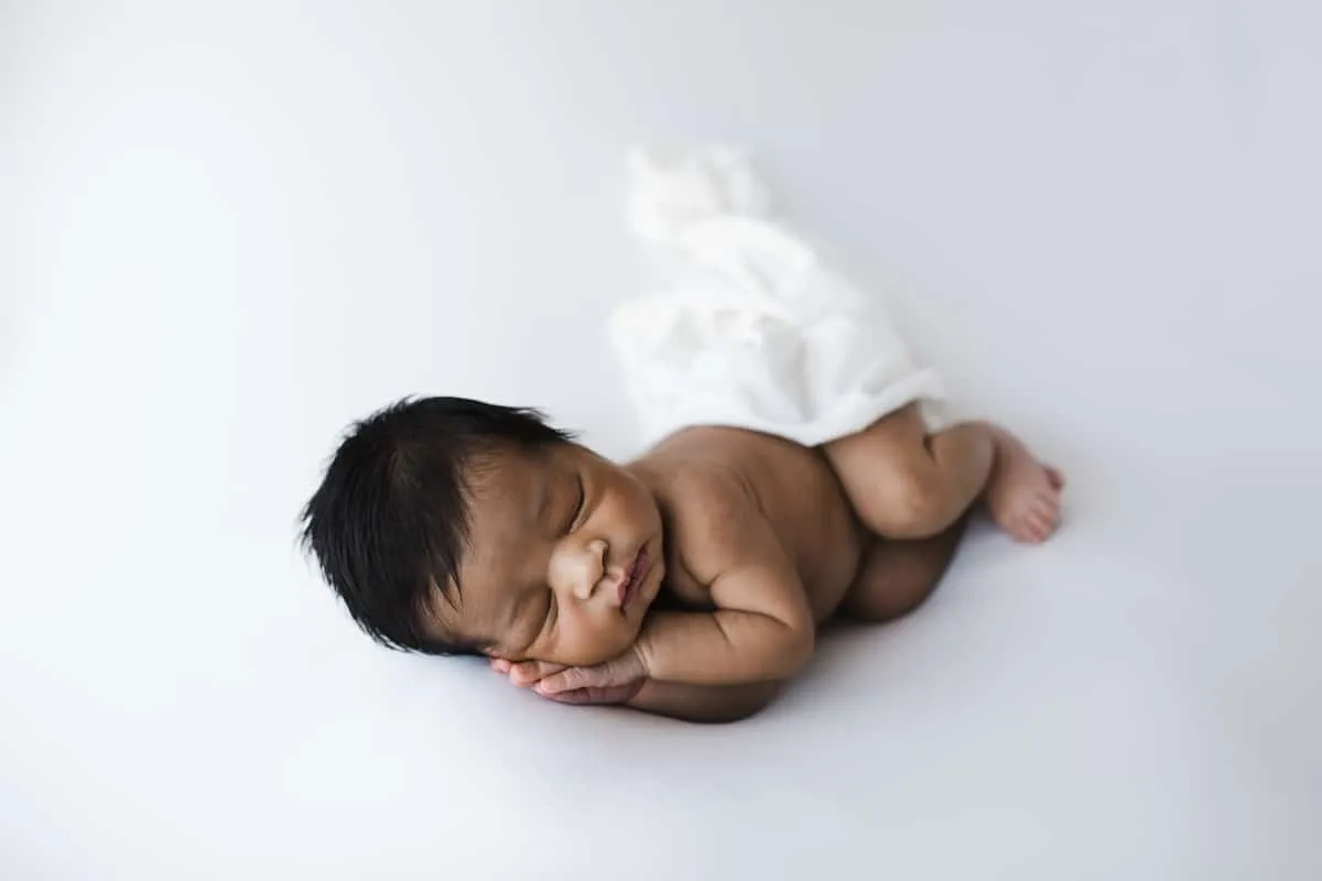 Newborn baby sleeping on a white surface with a white cloth partially covering their lower body, hands tucked under cheek.