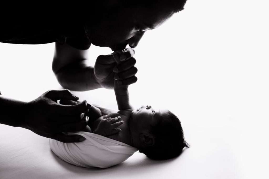 Adult kisses a newborn’s foot while gently holding the baby, who is swaddled in a white blanket and gazing upward. The image is in black and white with high contrast lighting.