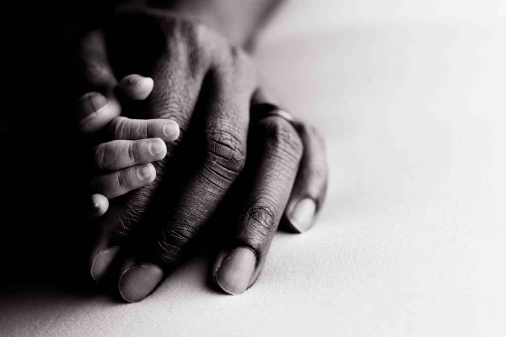 An adult hand gently rests on top of a baby's hand on a white surface, both hands partially in shadow.