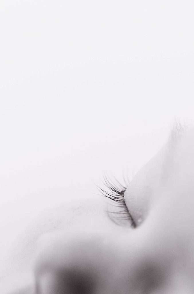 Close-up of a person's closed eye and eyelashes, photographed in black and white with a soft focus and minimal background.