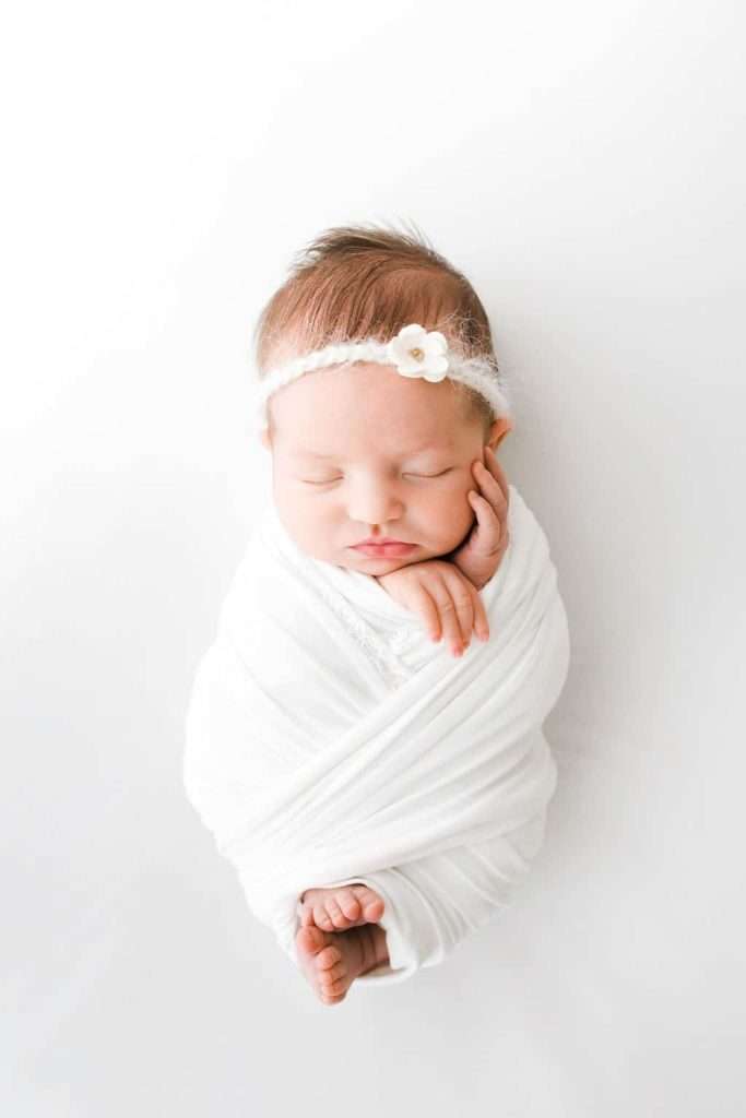 Newborn baby swaddled in white blanket, sleeping with hand on cheek, wearing a white headband with a small flower, on a white background.