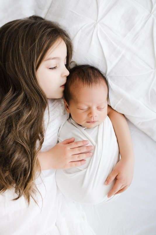 A young girl with long brown hair hugs a swaddled newborn baby while lying on a white bed, both looking peaceful and calm.