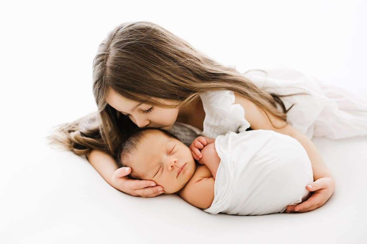 Young girl gently cradles and kisses a swaddled newborn baby on a white background.