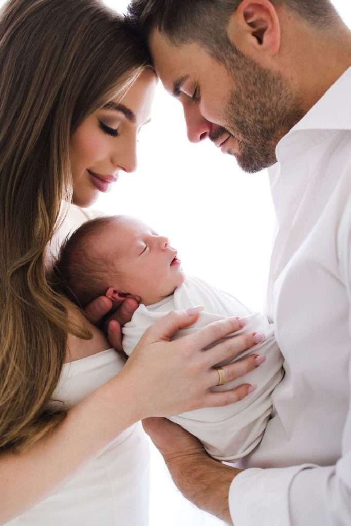 Man and woman gently cradle a newborn baby wrapped in a white blanket, foreheads touching, all dressed in white, against a bright background.
