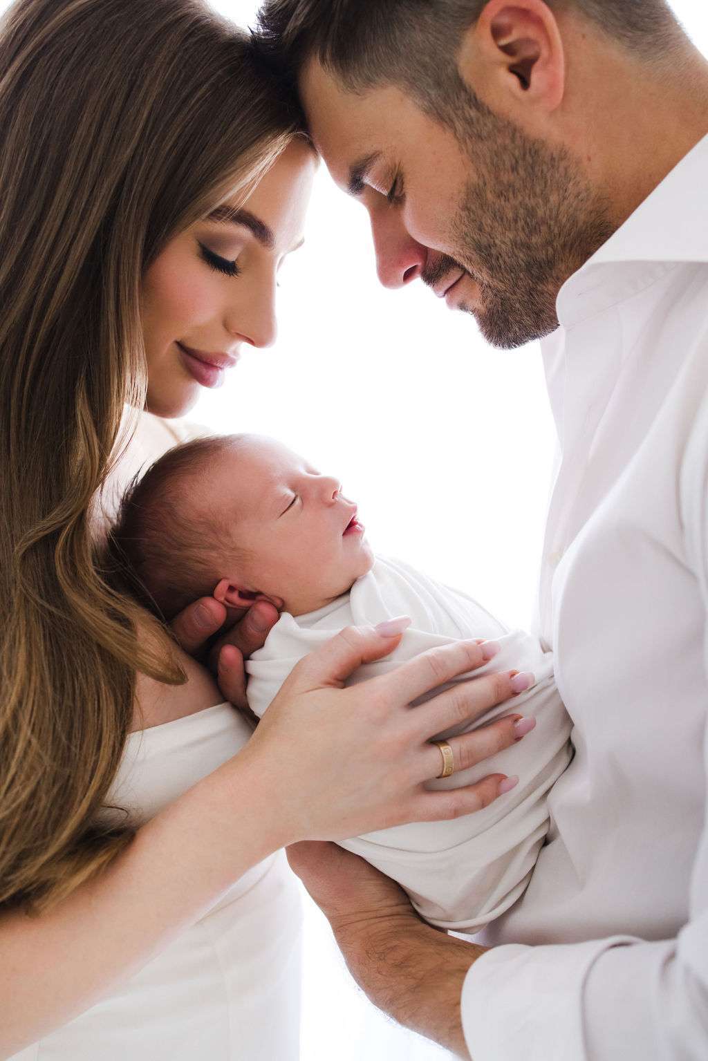 Man and woman gently cradle a newborn baby wrapped in a white blanket, foreheads touching, all dressed in white, against a bright background.
