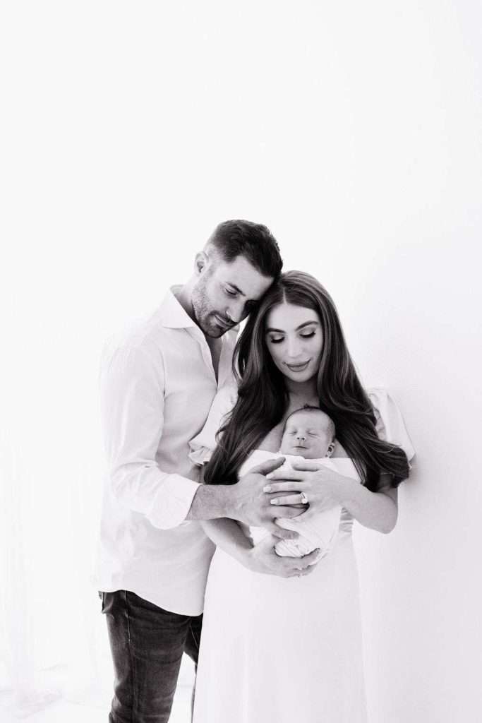A man and woman cradle a newborn baby together, standing close and looking down at the baby in a softly lit, white setting.