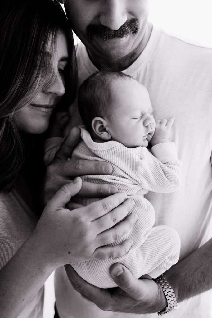 A woman and man gently hold a sleeping baby close to their chests, forming a close, intimate family portrait in black and white.