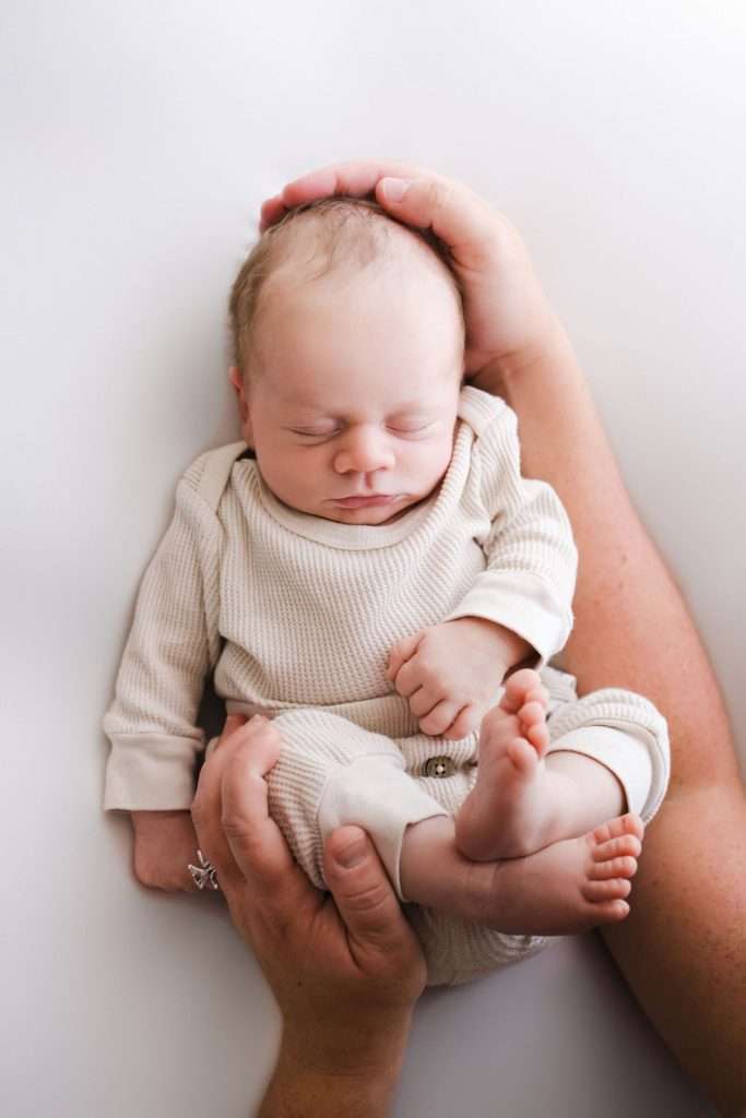 A newborn baby in a cream outfit sleeps peacefully while being gently cradled by adult hands on a white background.
