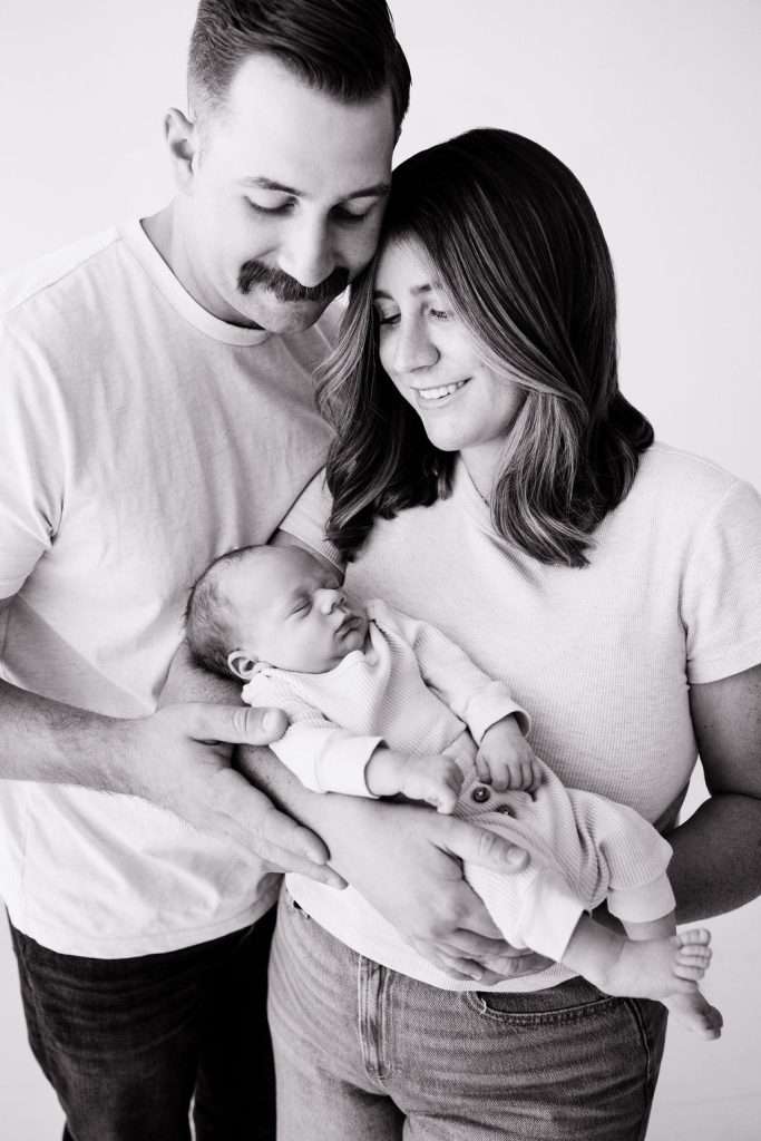 A man and woman smile while holding a sleeping baby in their arms. All three wear light-colored clothing. The image is in black and white.