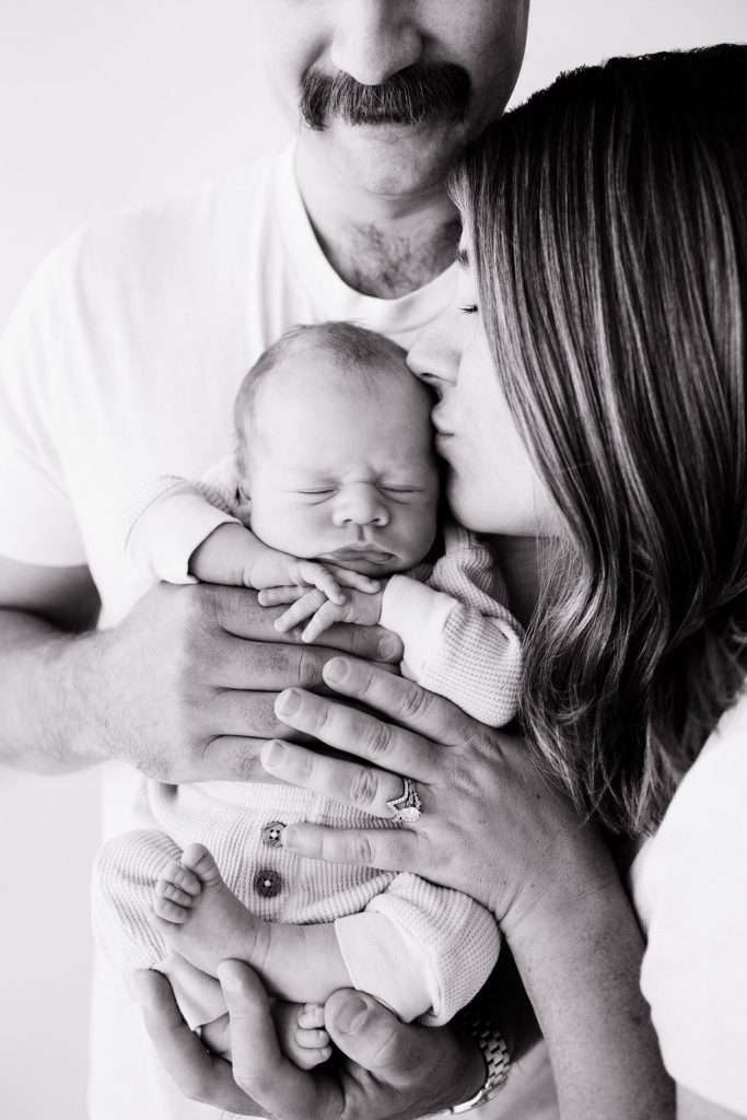 A woman kisses a newborn baby’s face while a man holds both of them from behind. All three are shown from the shoulders up in a close, intimate black-and-white portrait.