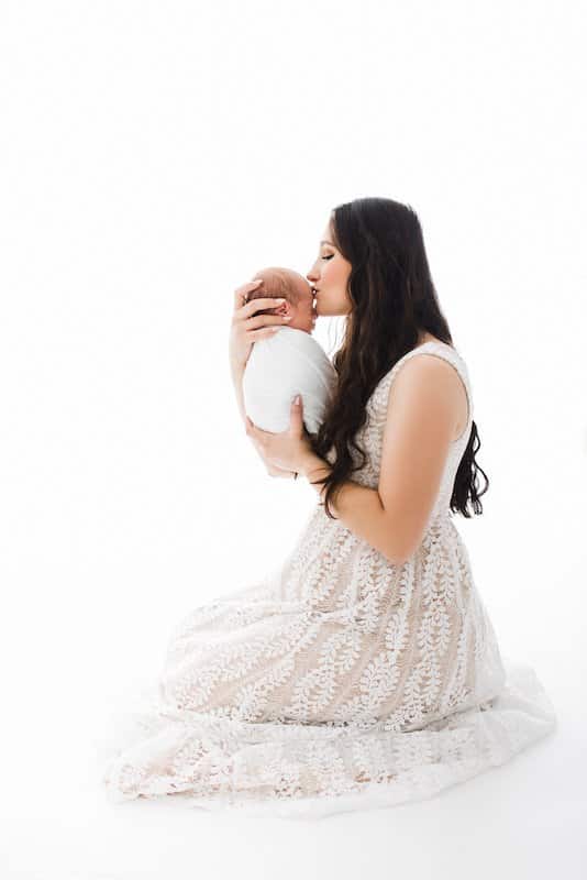 Woman in a white lace dress kneeling on the floor, holding and kissing a swaddled newborn baby against a white background.