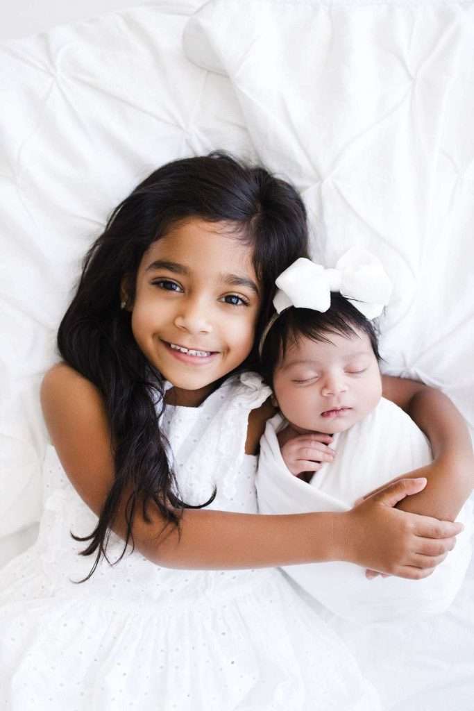 A young girl in a white dress hugs a swaddled newborn baby with a white bow on her head. Both are lying on a white bed.