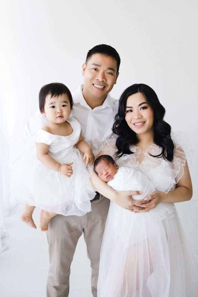 A family of four poses together against a white background. The parents smile, holding a toddler and a newborn, all dressed in white clothing.