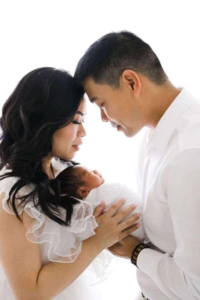 A couple stands close together, gently holding a newborn baby wrapped in a white blanket, all dressed in white against a bright background.