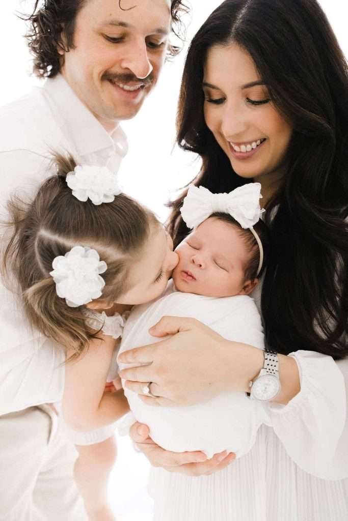 A smiling family of four, with a father, mother, young daughter kissing a newborn swaddled in white, all wearing white outfits and hair accessories.