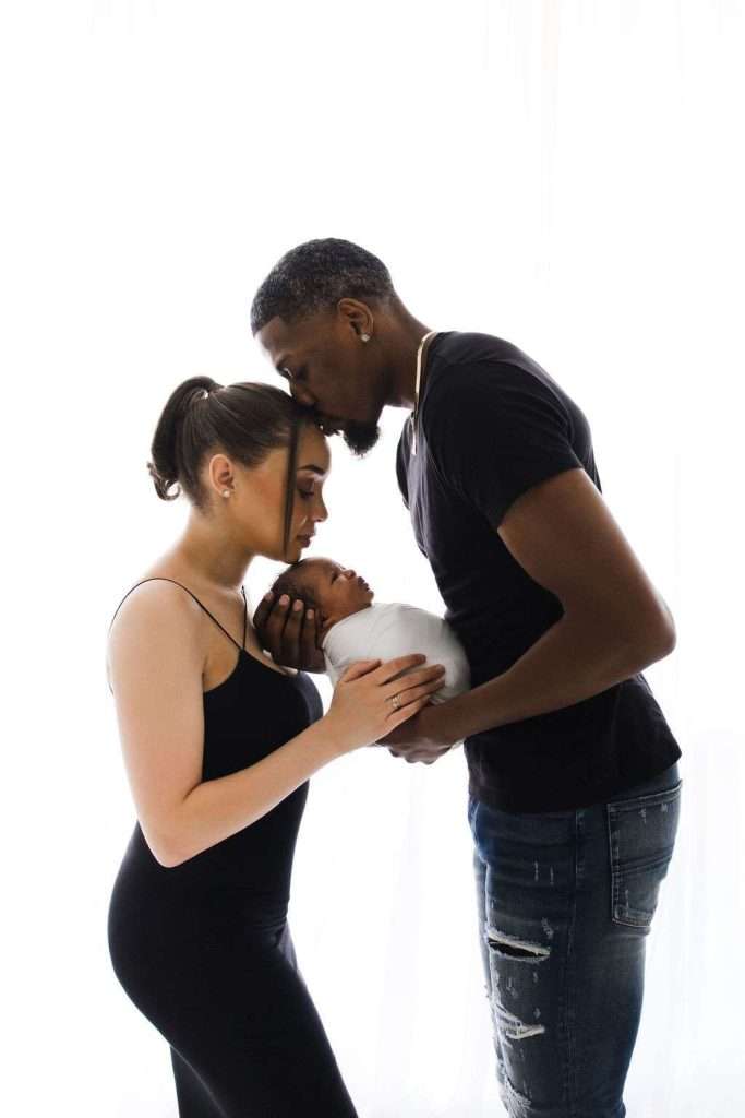 A man kisses a baby’s head while a woman holds the baby, both standing close together against a white background.