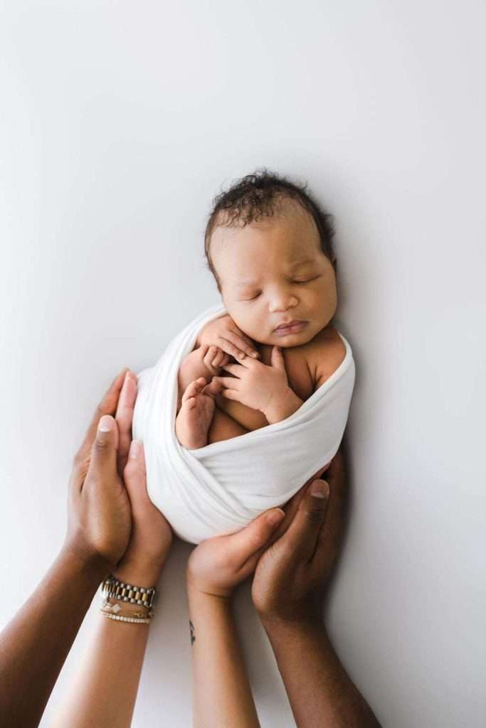 Newborn wrapped in a white blanket, sleeping peacefully, with adult hands gently cradling and supporting the baby from below against a white background.