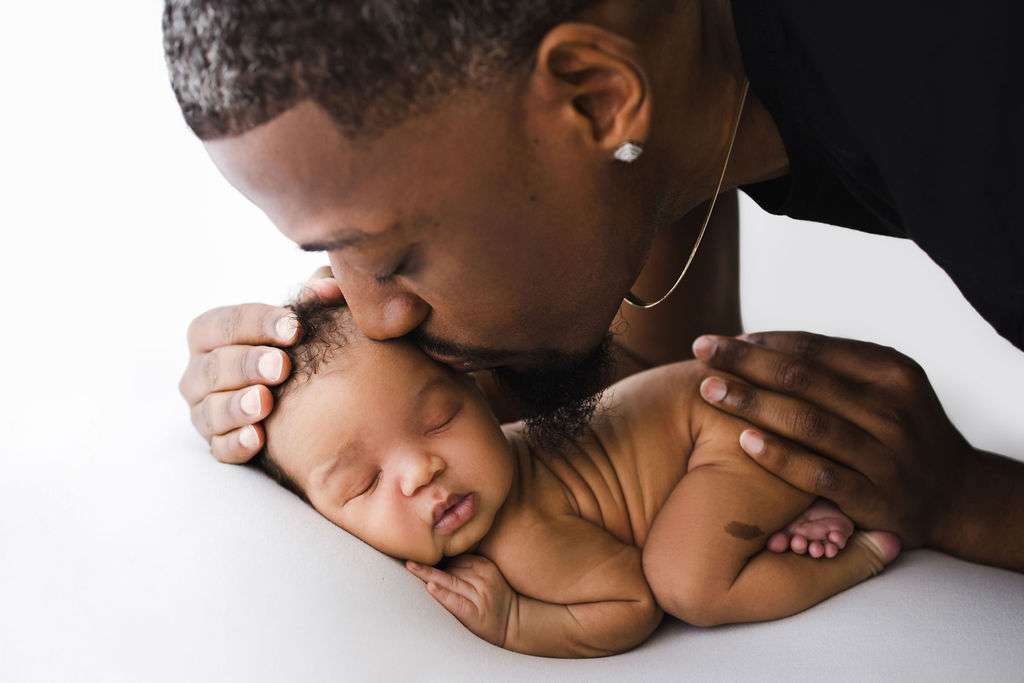 Man gently kisses a sleeping newborn baby, cradling the infant's head and back with his hands, against a white background.