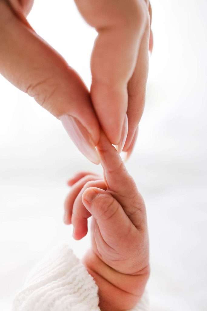 Close-up of an adult hand gently holding a newborn baby's fingers against a soft, light background.